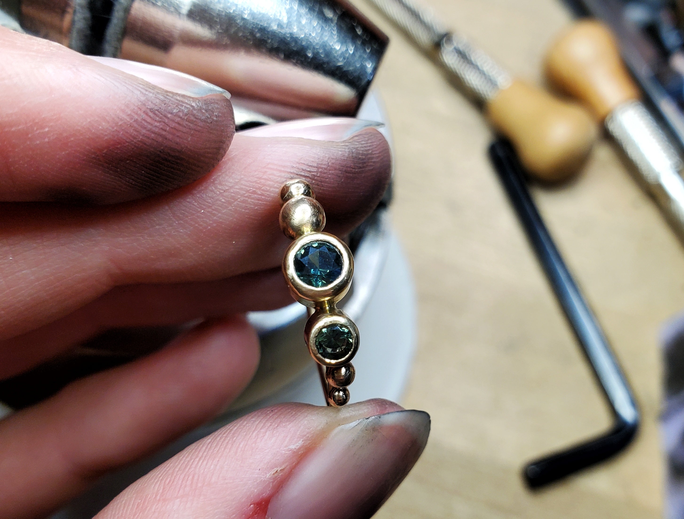 Hand holding an in-progress 14k gold ring with Madagascan sapphires over a bench of tools.